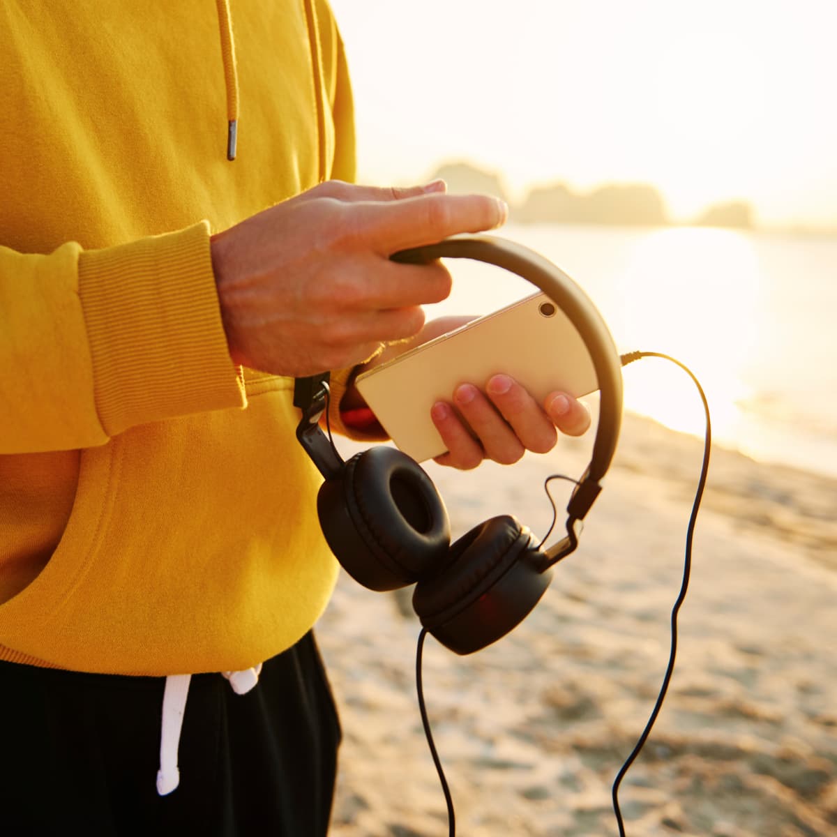 Hands holding a cell phone and headphones on the beach.